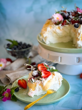 A beautifully presented dessert on a green plate, topped with fresh strawberries, blueberries, and mint leaves. There is a creamy texture with what appears to be dollops of whipped cream or possibly pavlova. The main dessert is displayed on a white stand in the background, partially sliced. Scattered around are flowers, adding a touch of elegance.