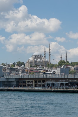 A large, historic mosque with multiple minarets rises amid a bustling cityscape. The foreground features a waterfront with various shops and restaurants, and people are visible walking on the pier. The sky is dotted with clouds, and the water appears calm and blue.