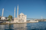 A waterfront mosque with two tall minarets stands prominently next to the sea, with a large suspension bridge in the background. A ferry moves along the water, and people are gathered near the shore, with trees and buildings surrounding the scene.