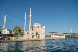 A waterfront mosque with two tall minarets stands prominently next to the sea, with a large suspension bridge in the background. A ferry moves along the water, and people are gathered near the shore, with trees and buildings surrounding the scene.