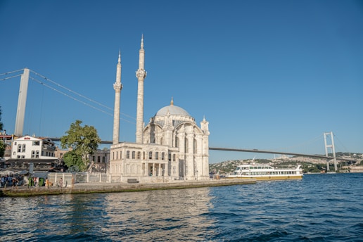 A waterfront mosque with two tall minarets stands prominently next to the sea, with a large suspension bridge in the background. A ferry moves along the water, and people are gathered near the shore, with trees and buildings surrounding the scene.