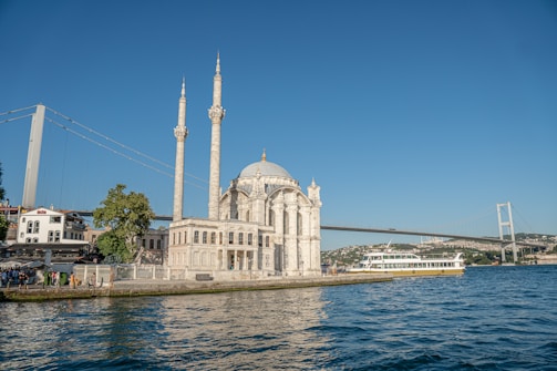 A waterfront mosque with two tall minarets stands prominently next to the sea, with a large suspension bridge in the background. A ferry moves along the water, and people are gathered near the shore, with trees and buildings surrounding the scene.