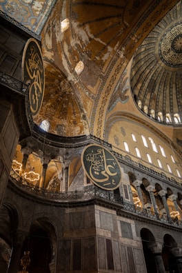 Intricate and grand architectural details are visible, emphasizing ornate arabesque designs and large, circular calligraphy medallions hanging on the walls. Towering arches and domed ceilings adorned with mosaic patterns evoke a sense of historical and cultural richness.