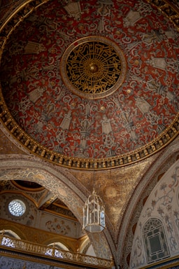 An intricately decorated dome featuring detailed patterns and Arabic calligraphy, with vibrant red, gold, and black colors. The surrounding architecture includes ornate arches and a hanging lantern, showcasing a rich mix of floral and geometric designs.