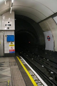 An underground subway station platform with visible safety markings on the floor and a dark tunnel entrance. The platform has warning signs about high voltage and a cautionary sign reminding passengers to mind the gap. The station includes lighting fixtures, tiled walls, and a distinctive logo on the wall indicating the station's location.