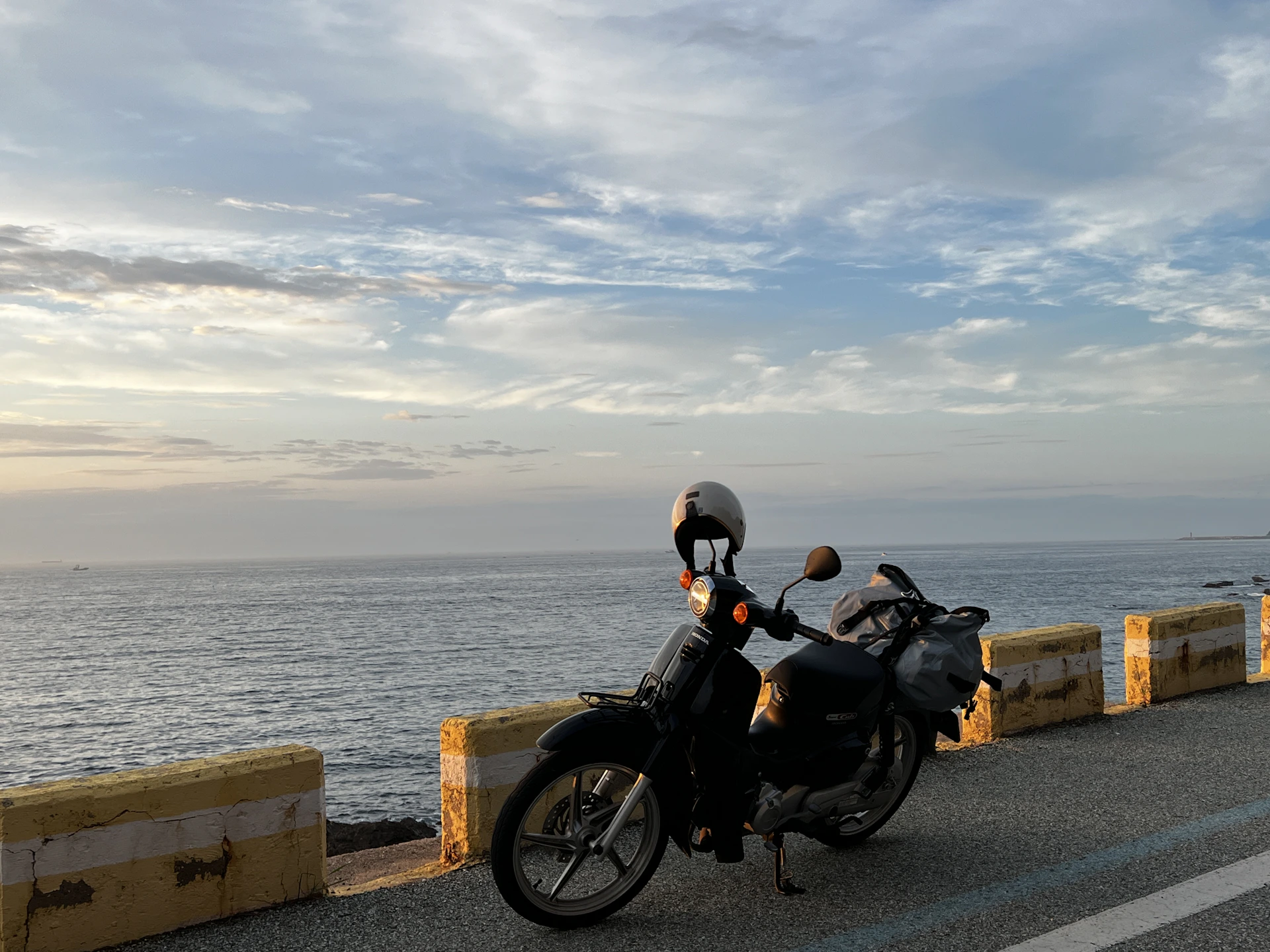 A rider enjoying a scenic coastal road on a Honda bike, with waves crashing nearby and clear blue skies overhead.