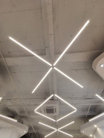 Geometric LED light fixtures are mounted on a white industrial-style ceiling, with an air conditioning unit visible near the center. The lights form intersecting diagonal lines, creating a modern and minimalistic design.