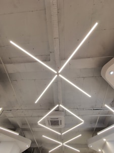Geometric LED light fixtures are mounted on a white industrial-style ceiling, with an air conditioning unit visible near the center. The lights form intersecting diagonal lines, creating a modern and minimalistic design.