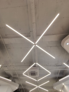 Geometric LED light fixtures are mounted on a white industrial-style ceiling, with an air conditioning unit visible near the center. The lights form intersecting diagonal lines, creating a modern and minimalistic design.