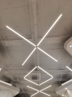 Geometric LED light fixtures are mounted on a white industrial-style ceiling, with an air conditioning unit visible near the center. The lights form intersecting diagonal lines, creating a modern and minimalistic design.
