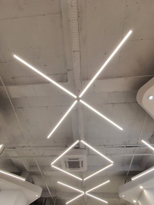 Geometric LED light fixtures are mounted on a white industrial-style ceiling, with an air conditioning unit visible near the center. The lights form intersecting diagonal lines, creating a modern and minimalistic design.