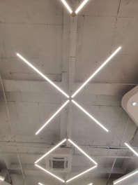 A modern ceiling design featuring intersecting linear light fixtures forming an 'X' shape. The structure includes an air conditioning unit set within a minimalist and industrial setting, marked by a bare and slightly rough ceiling finish.