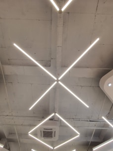 A modern ceiling design featuring intersecting linear light fixtures forming an 'X' shape. The structure includes an air conditioning unit set within a minimalist and industrial setting, marked by a bare and slightly rough ceiling finish.