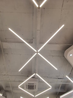 A modern ceiling design featuring intersecting linear light fixtures forming an 'X' shape. The structure includes an air conditioning unit set within a minimalist and industrial setting, marked by a bare and slightly rough ceiling finish.