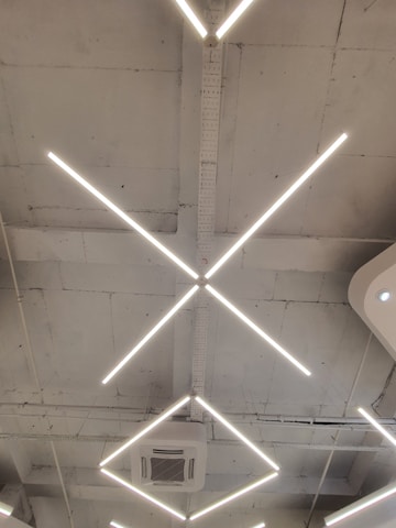 A modern ceiling design featuring intersecting linear light fixtures forming an 'X' shape. The structure includes an air conditioning unit set within a minimalist and industrial setting, marked by a bare and slightly rough ceiling finish.