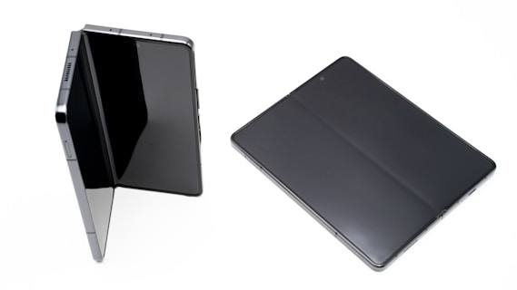 Two electronic devices are shown, one in a partially open position and the other lying flat on a white background. Both devices have large, black touchscreens.