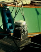 A close-up of a mooring setup for a boat. The main focus is on a weathered wooden bollard with a white cap at the top, which is securing several ropes. A blue boat fender is seen hanging from the side of the boat, which has a green hull and wooden trim. The deck of the boat, featuring wooden elements and seating, can be observed partially.