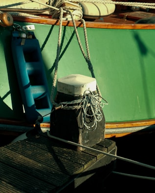 A close-up of a mooring setup for a boat. The main focus is on a weathered wooden bollard with a white cap at the top, which is securing several ropes. A blue boat fender is seen hanging from the side of the boat, which has a green hull and wooden trim. The deck of the boat, featuring wooden elements and seating, can be observed partially.