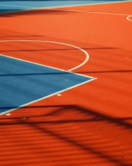 A vibrant sports court with sections of red and blue, featuring crisp white lines marking the boundaries. Shadows from a structure create geometric patterns across the surface, adding depth and interest to the scene.