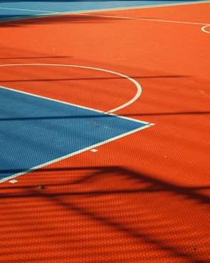 A vibrant sports court with sections of red and blue, featuring crisp white lines marking the boundaries. Shadows from a structure create geometric patterns across the surface, adding depth and interest to the scene.