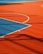A vibrant sports court with sections of red and blue, featuring crisp white lines marking the boundaries. Shadows from a structure create geometric patterns across the surface, adding depth and interest to the scene.