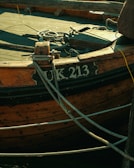 A close-up view of an old wooden boat with the registration number UK 213. The boat has weathered wood planks and is secured with several ropes tied to its structure.