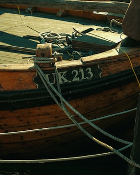 A close-up view of an old wooden boat with the registration number UK 213. The boat has weathered wood planks and is secured with several ropes tied to its structure.
