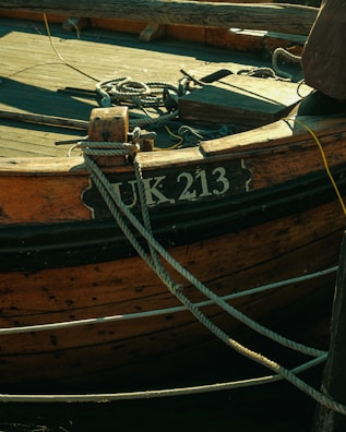 A close-up view of an old wooden boat with the registration number UK 213. The boat has weathered wood planks and is secured with several ropes tied to its structure.