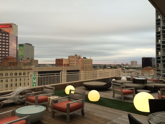 A rooftop terrace with modern outdoor furniture, featuring wooden lounge chairs with red cushions and circular glowing lights. The background consists of a cityscape with mid-rise and high-rise buildings under a cloudy sky.