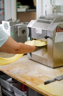 A person is operating a pasta machine, feeding dough through rollers on a wooden table. The setting appears to be a kitchen or food preparation area with various equipment visible in the background.