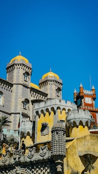 A castle with elaborate architectural details featuring yellow domes and intricate stone carvings. The building has a mix of styles, including Gothic and Moorish influences, with a vibrant blue sky as a backdrop. People are seen standing near the stone railing, adding a sense of scale to the structure.