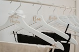 Several white hangers on a rack, each customized with cursive names in gold lettering. Some hangers hold black and beige garments. One hanger is adorned with a decorative white bow featuring a small jewel.