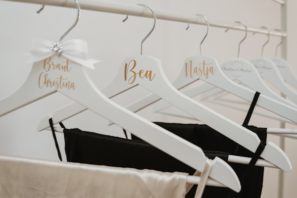 Several white hangers on a rack, each customized with cursive names in gold lettering. Some hangers hold black and beige garments. One hanger is adorned with a decorative white bow featuring a small jewel.