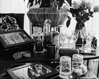 A luxuriously arranged setting featuring glasses, decanters, a cigar humidor, and vases of roses. Crystal glassware is neatly lined on a tray alongside bottled drinks, and a pair of sunglasses rests on the table. The scene is elegantly composed with a combination of modern and classic touches, exuding a sense of sophistication and refinement.