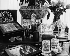 A luxuriously arranged setting featuring glasses, decanters, a cigar humidor, and vases of roses. Crystal glassware is neatly lined on a tray alongside bottled drinks, and a pair of sunglasses rests on the table. The scene is elegantly composed with a combination of modern and classic touches, exuding a sense of sophistication and refinement.