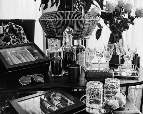 A luxuriously arranged setting featuring glasses, decanters, a cigar humidor, and vases of roses. Crystal glassware is neatly lined on a tray alongside bottled drinks, and a pair of sunglasses rests on the table. The scene is elegantly composed with a combination of modern and classic touches, exuding a sense of sophistication and refinement.