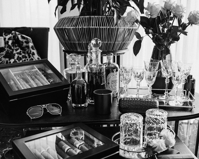 A luxuriously arranged setting featuring glasses, decanters, a cigar humidor, and vases of roses. Crystal glassware is neatly lined on a tray alongside bottled drinks, and a pair of sunglasses rests on the table. The scene is elegantly composed with a combination of modern and classic touches, exuding a sense of sophistication and refinement.