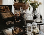 A collection of luxury items including crystal glasses, liquor bottles, and a wooden cigar box arranged on a dark tabletop. A pair of sunglasses and a designer case are also present. In the background, a bouquet of white roses in a vase adds an elegant touch.