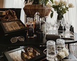 A collection of luxury items including crystal glasses, liquor bottles, and a wooden cigar box arranged on a dark tabletop. A pair of sunglasses and a designer case are also present. In the background, a bouquet of white roses in a vase adds an elegant touch.
