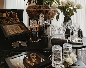 A collection of luxury items including crystal glasses, liquor bottles, and a wooden cigar box arranged on a dark tabletop. A pair of sunglasses and a designer case are also present. In the background, a bouquet of white roses in a vase adds an elegant touch.