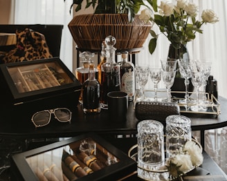 A collection of luxury items including crystal glasses, liquor bottles, and a wooden cigar box arranged on a dark tabletop. A pair of sunglasses and a designer case are also present. In the background, a bouquet of white roses in a vase adds an elegant touch.