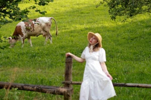 A peaceful rural scene with a cow grazing on lush green grass. In the foreground, a woman in a white dress and wide-brimmed hat stands by a wooden fence, surrounded by trees.