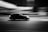 A black and white photograph capturing a car moving at high speed. The background appears blurred, emphasizing the motion of the car. The scene is set at night, with lights streaking across the sky, adding to the dynamic feel.