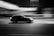 A black and white photograph capturing a car moving at high speed. The background appears blurred, emphasizing the motion of the car. The scene is set at night, with lights streaking across the sky, adding to the dynamic feel.