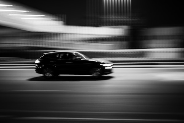 A black and white photograph capturing a car moving at high speed. The background appears blurred, emphasizing the motion of the car. The scene is set at night, with lights streaking across the sky, adding to the dynamic feel.