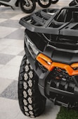 A close-up view of an all-terrain vehicle with a large, rugged tire featuring a distinct tread pattern. The vehicle's front features a bold orange accent on its matte black body, showcasing its robust and sturdy build. In the background, other vehicles and parts of their wheels are visible, softly blurred.