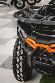 A close-up view of an all-terrain vehicle with a large, rugged tire featuring a distinct tread pattern. The vehicle's front features a bold orange accent on its matte black body, showcasing its robust and sturdy build. In the background, other vehicles and parts of their wheels are visible, softly blurred.