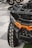 A close-up view of an all-terrain vehicle with a large, rugged tire featuring a distinct tread pattern. The vehicle's front features a bold orange accent on its matte black body, showcasing its robust and sturdy build. In the background, other vehicles and parts of their wheels are visible, softly blurred.