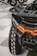 A close-up view of an all-terrain vehicle with a large, rugged tire featuring a distinct tread pattern. The vehicle's front features a bold orange accent on its matte black body, showcasing its robust and sturdy build. In the background, other vehicles and parts of their wheels are visible, softly blurred.