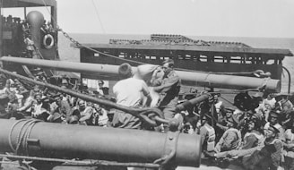 A crowded deck of a ship features numerous men dressed in military uniforms, some of whom are sitting and others standing. Two of these men are engaged in what appears to be a task or activity involving a large piece of machinery or equipment. The sea is visible in the background, indicating that the ship is at sea.